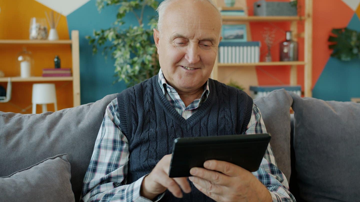 Starszy mężczyzna uśmiecha się, korzystając z tabletu cyfrowego na kanapie.